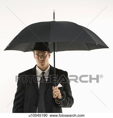 Stock Image - Caucasian mid-adult businessman wearing fedora holding umbrella and looking at viewer.. Fotosearch