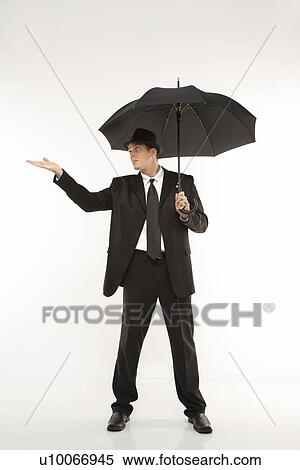 Stock Photography - Caucasian mid-adult businessman wearing fedora holding umbrella with arm outstretched.. Fotosearch