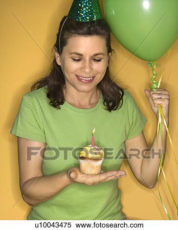 Stock Photography - Caucasian prime adult female holding cupcake with lit candle wearing party hat.. Fotosearch