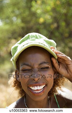 Stock Image - MId-adult Black female smiling with hand on cap and squinted eyes.. Fotosearch