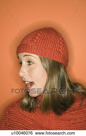 Stock Photograph - Side view of young adult Caucasian woman on orange background wearing winter hat and scarf.. Fotosearch
