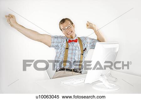 Stock Photography - Caucasian young man dressed like nerd stretching arms out and happy in front of computer.. Fotosearch