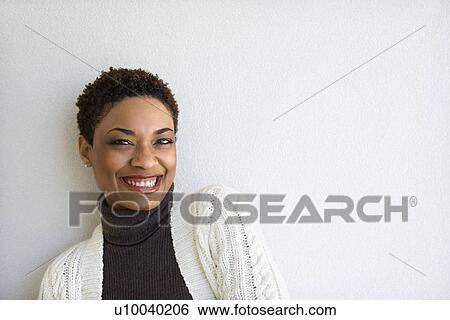 Stock Photograph - Close up of African- American woman standing against white wall looking at viewer and smiling.. Fotosearch