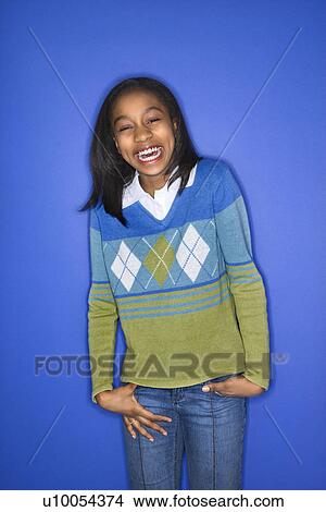 Picture - Portrait of African-American girl with hands in her pockets smiling standing against blue background.. Fotosearch
