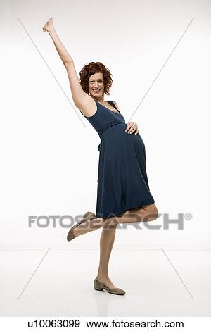 Stock Photo - Portrait of Caucasion mid-adult attractive pregnant woman standing with one knee up, one hand on belly and other arm in air looking at viewer and smiling.. Fotosearch
