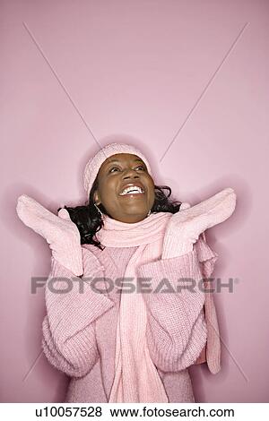 Stock Photo - Portrait of smiling African-American mid-adult woman on pink background wearing winter coat, hat, and scarf looking upward.. Fotosearch