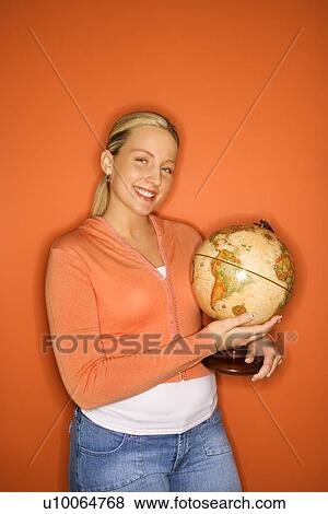 Stock Photo - Portrait of smiling blond Caucasian teen girl holding globe standing against orange background.. Fotosearch