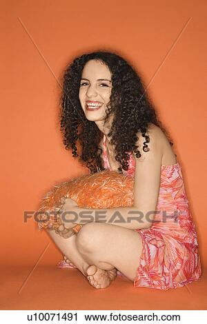 Stock Image - Smiling young Caucasian woman sitting on floor with pillow on orange background.. Fotosearch
