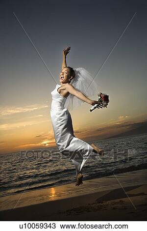 Stock Image - Young adult female Caucasian bride jumping on beach.. Fotosearch