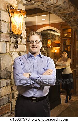 Stock Photograph - Caucasian man standing with arms crossed looking at viewer and smiling with Caucasian women in background with laptop.. Fotosearch