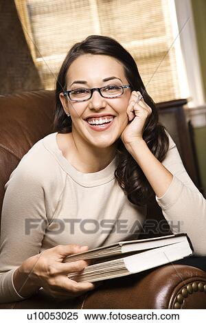 Stock Photography - Caucasian/Hispanic young woman sitting in leather chair holding book and smiling at viewer.. Fotosearch
