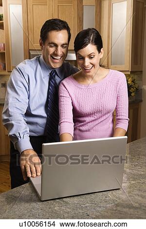 Mid-adult couple in kitchen looking and smiling at laptop computer. View Large Photo Image Picture - Mid-adult couple in kitchen looking and smiling at laptop computer.. Fotosearch