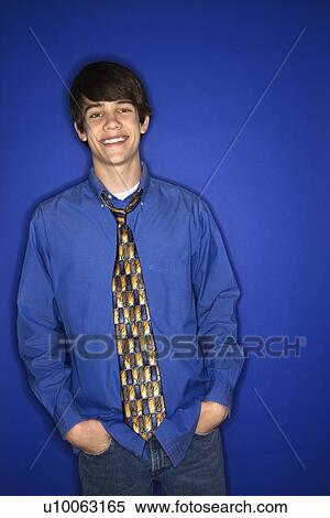 Stock Photography - Portrait of Caucasian teen boy standing with hands in pockets against blue background.. Fotosearch