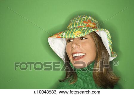 Stock Image - Portrait of smiling young adult Caucasian woman on green background wearing floppy hat.. Fotosearch