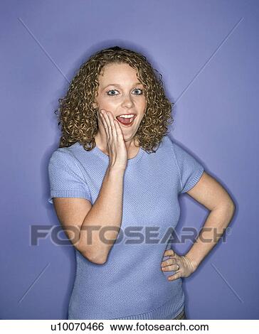 Stock Photograph - Portrait of young adult Caucasian woman on blue background looking surprised.. Fotosearch