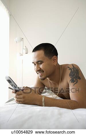 Stock Photo - Smiling shirtless Asian young man lying on bed dialing or text messaging on cellphone.. Fotosearch
