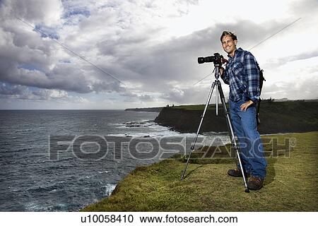Stock Image - Caucasian mid-adult male standing with camera on tripod looking at viewer on cliff overlooking the ocean in Maui, Hawaii.. Fotosearch