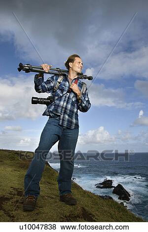 Stock Photo - Caucasian mid-adult man standing with camera and tripod over his shoulder on cliff overlooking ocean in Maui, Hawaii.. Fotosearch