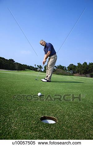 Stock Photography - Caucasion mid-adult man putting golf ball with hole in foreground.. Fotosearch