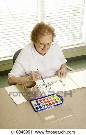 Stock Image - Elderly Caucasian woman painting with watercolors at retirement community center.. Fotosearch