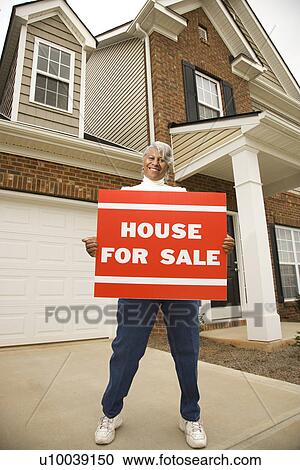Stock Image - Portrait of middle-aged African-American female outside house with for sale sign.. Fotosearch
