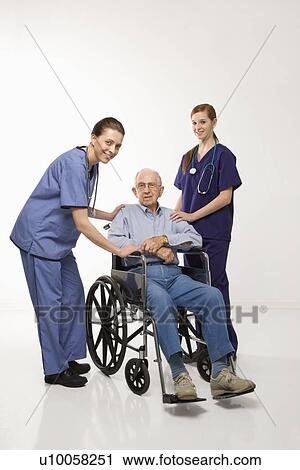 Stock Image - Two Caucasian females wearing scrubs with elderly Caucasian male in wheelchair.. Fotosearch