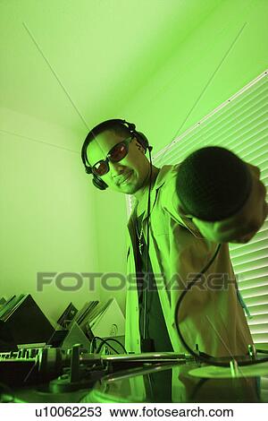 Stock Image - Asian young adult male DJ standing behind mixing equipment and turntable holding microphone out to viewer.. Fotosearch