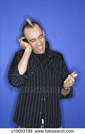 Stock Photo - Caucasian man smiling with mohawk listening to headphones holding cd standing against blue background.. Fotosearch