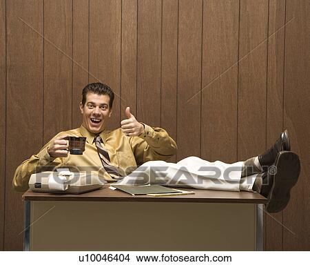 Picture - Caucasion mid-adult retro businessman sitting with feet propped on desk drinking coffee giving a thumbs up.. Fotosearch