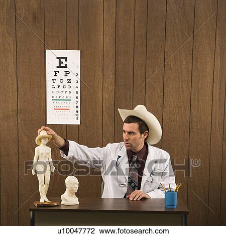 Stock Image - Mid-adult Caucasian male doctor wearing cowboy hat sitting at desk playing with figurine.. Fotosearch