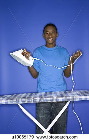 Portrait of smiling African-American teen boy holding iron and cord standing in behind ironing board against blue background. View Large Photo Image Stock Photo - Portrait of smiling African-American teen boy holding iron and cord standing in behind ironing board against blue background.. Fotosearch