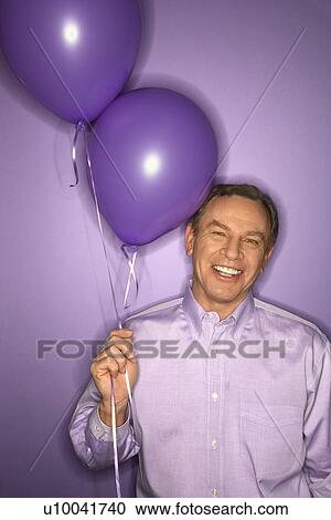 Stock Image - Smiling middle-aged Caucasian man on purple background holding purple balloons.. Fotosearch
