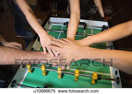 Stock Photography - Group of young friends clasping hands in solidarity over foosball table.. Fotosearch