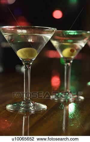 Stock Image - Two martinis with olives on bar with red and green lights in background.. Fotosearch