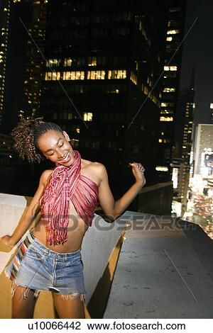 Stock Image - Young sexy African-American woman jumping and screaming with excitement on patio in New York City, New York at night.. Fotosearch
