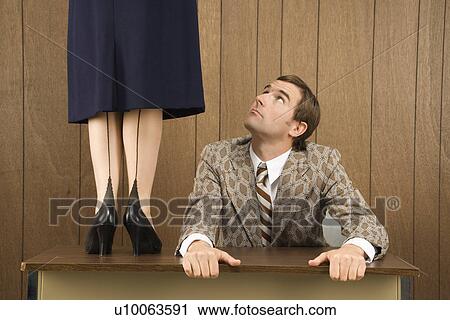 Stock Image - Mid-adult Caucasian male holding desk looking up to Caucasian female standing on desk.. Fotosearch