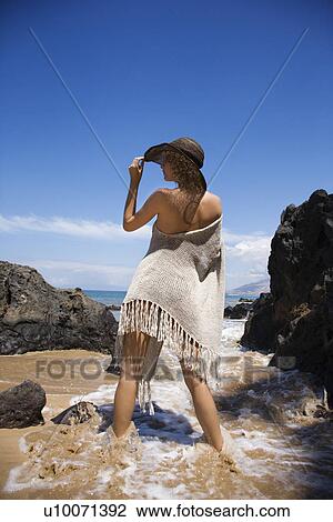 Stock Image - Young adult female Caucasian standing in water on beach tilting brimmed hat.. Fotosearch