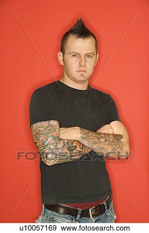 Stock Photo - Caucasian man with mohawk and tattoos standing with arms crossed against orange background.. Fotosearch