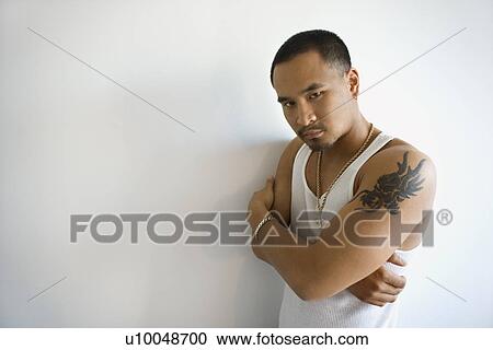 Stock Image - Portrait of Asian young adult man leaning against white wall with arms crossed looking at viewer.. Fotosearch