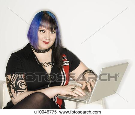Stock Photography - Portrait of Caucasian woman with blue hair, tattoo, and spike collar typing on laptop against white background.. Fotosearch