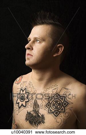 Stock Image - Portrait of shirtless Caucasian man with tattoos and mohawk against black background.. Fotosearch
