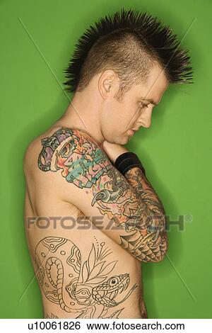 Stock Photograph - Profile of Caucasian man with mohawk and tattoos standing against green background.. Fotosearch