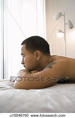 Stock Image - Profile of shirtless Asian young adult man lying on bed resting head on arms looking out window.. Fotosearch