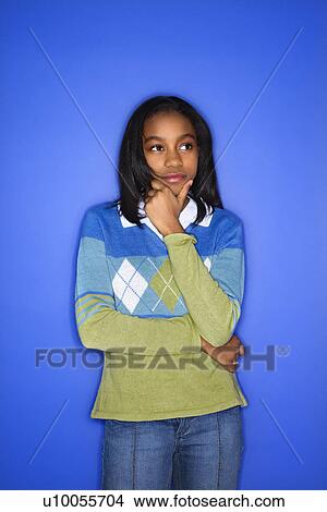 Picture - Portrait of African-American girl with hand on chin standing in front of blue background.. Fotosearch
