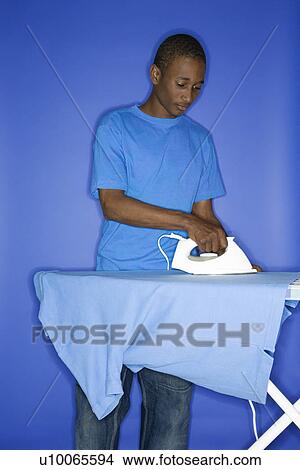 Picture - Portrait of African-American teen boy ironing shirt standing against blue background.. Fotosearch