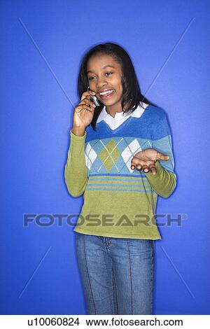 Portrait of African-American teen girl smiling and talking on cellphone standing in front of blue background. View Large Photo Image Picture - Portrait of African-American teen girl smiling and talking on cellphone standing in front of blue background.. Fotosearch