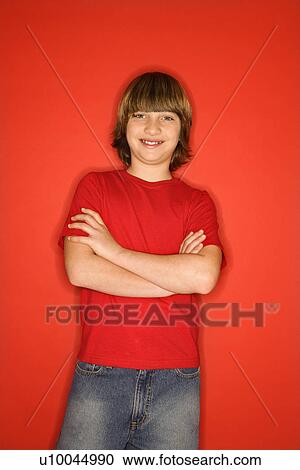 Stock Image - Portrait of Caucasian boy in studio standing against red background with arms crossed.. Fotosearch