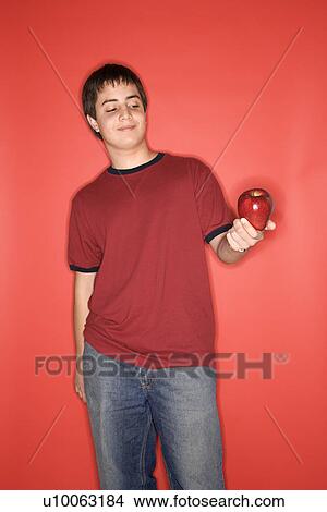 Portrait of Caucasian teen boy holding an apple standing against red background. View Large Photo Image Picture - Portrait of Caucasian teen boy holding an apple standing against red background.. Fotosearch