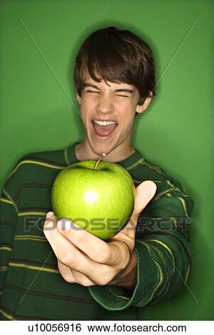 Stock Photograph - Portrait of Caucasian teen boy holding apple out in hand and making facial expression.. Fotosearch