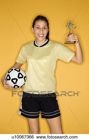 Stock Photograph - Portrait of Multi-racial teen girl holding soccer ball and trophy smiling standing against yellow background.. Fotosearch